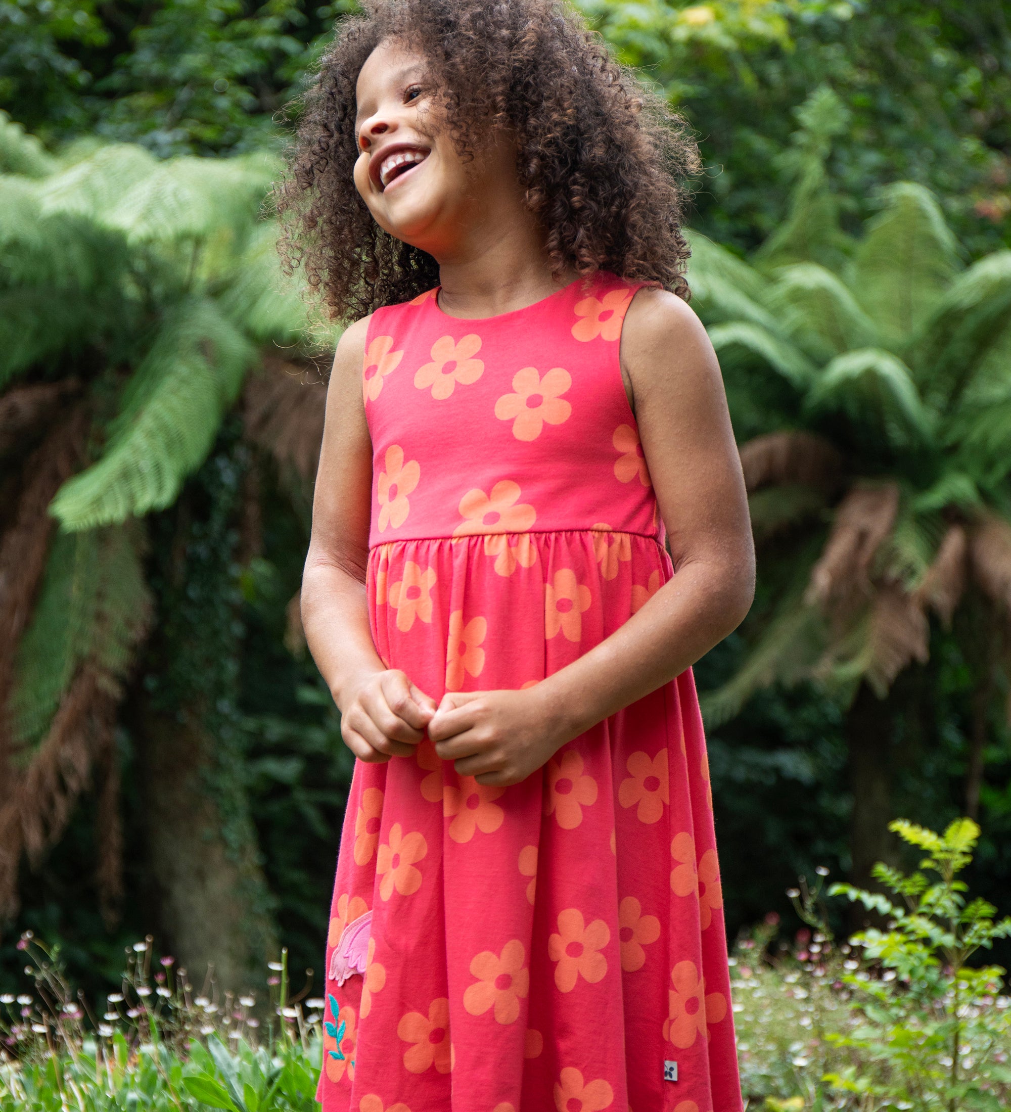 A child standing on grass outside wearing the Frugi Samantha summer dress - a light pink colour with a orange floral print and a flamingo appliqué 