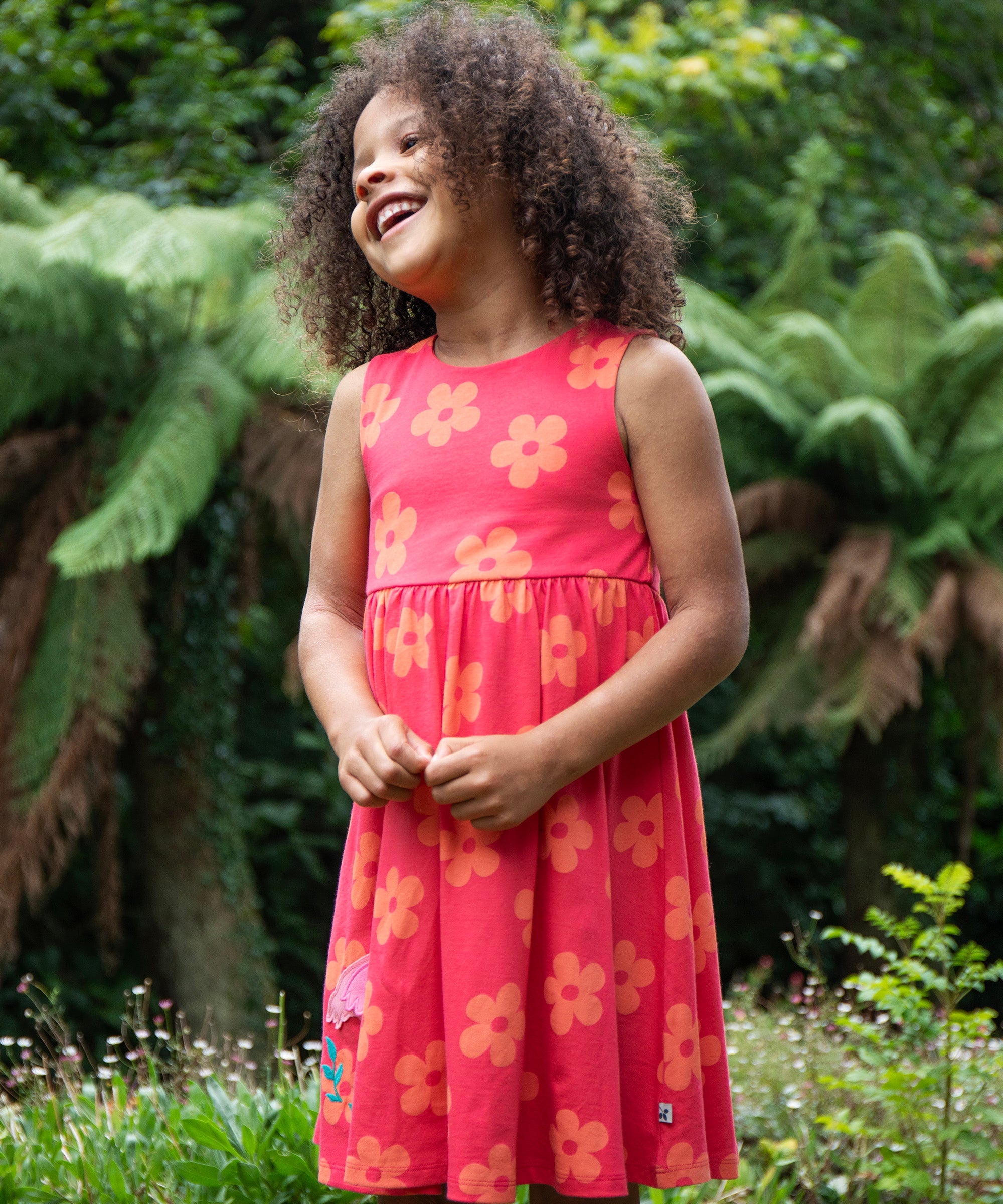 A child standing on grass outside wearing the Frugi Samantha summer dress - a light pink colour with a orange floral print and a flamingo appliqué 