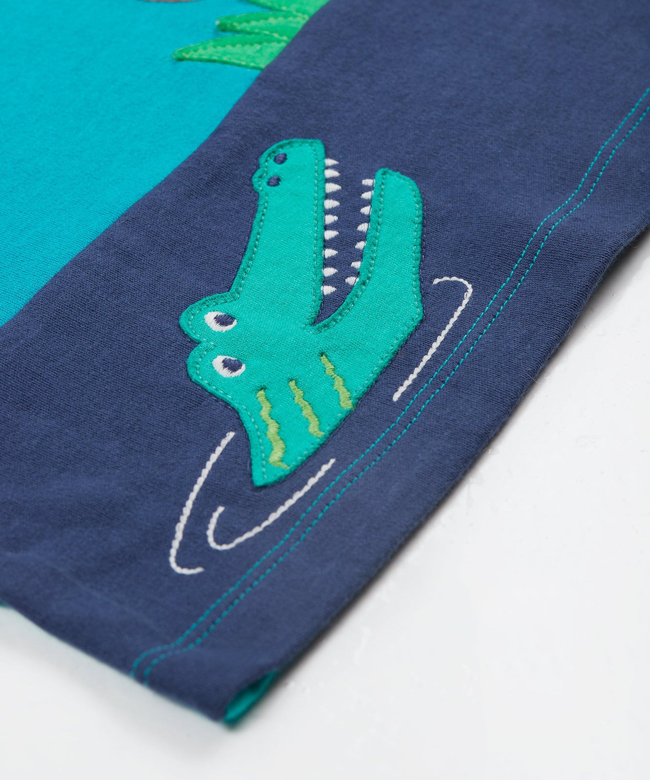 A close up of the Frugi sea, land, canopy short sleeve -shirt. Showing the crocodil applique and light blue stitching on the hem of the t-shirt on blue organic cotton fabric