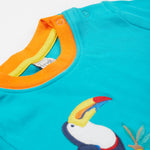 A close up of the Frugi sea, land, canopy short sleeve -shirt. Showing the shoulder fastener, orange neck trim and the toucan applique stitched on a blue organic cotton fabric