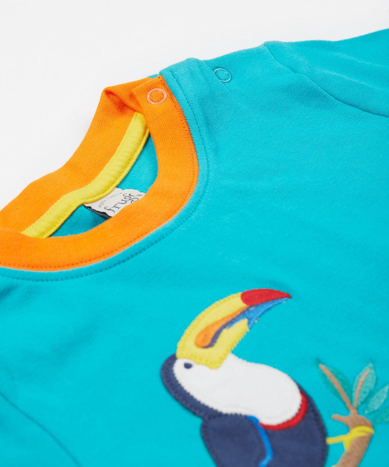 A close up of the Frugi sea, land, canopy short sleeve -shirt. Showing the shoulder fastener, orange neck trim and the toucan applique stitched on a blue organic cotton fabric