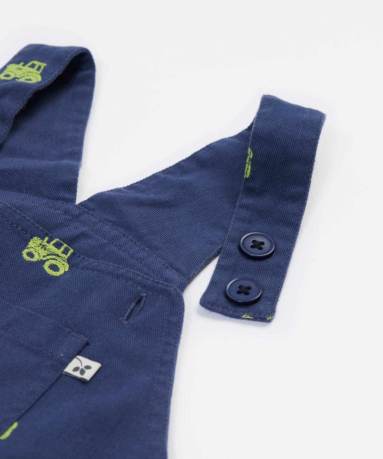 Close up of the Frugi baby short navy tractor dungarees. Showing the button fastener detail and green tractor embroidery on navy organic cotton fabric.