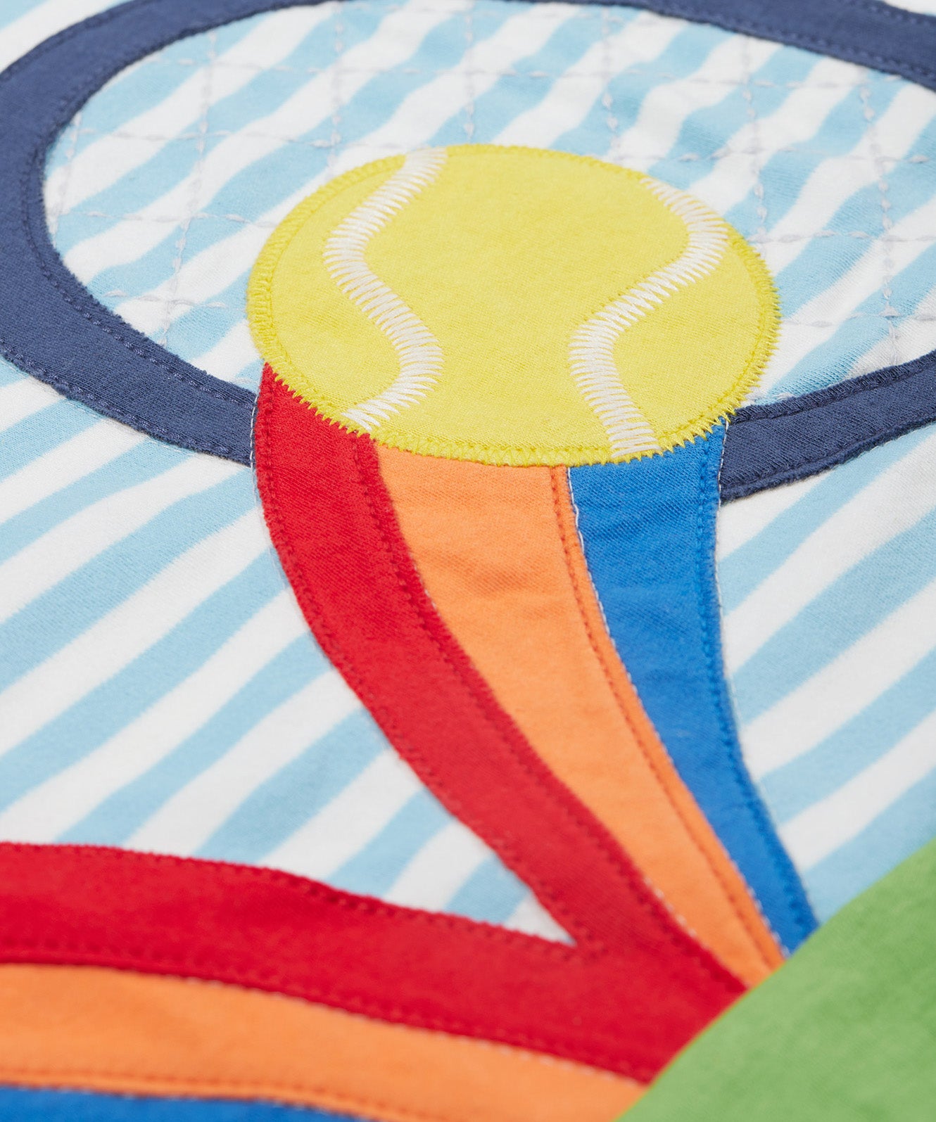 A close up of the Frugi kids bluebird tennis t-shirt. Showing the tennis racket, tennis ball and rainbow appliqué on blue and white striped organic cotton fabric