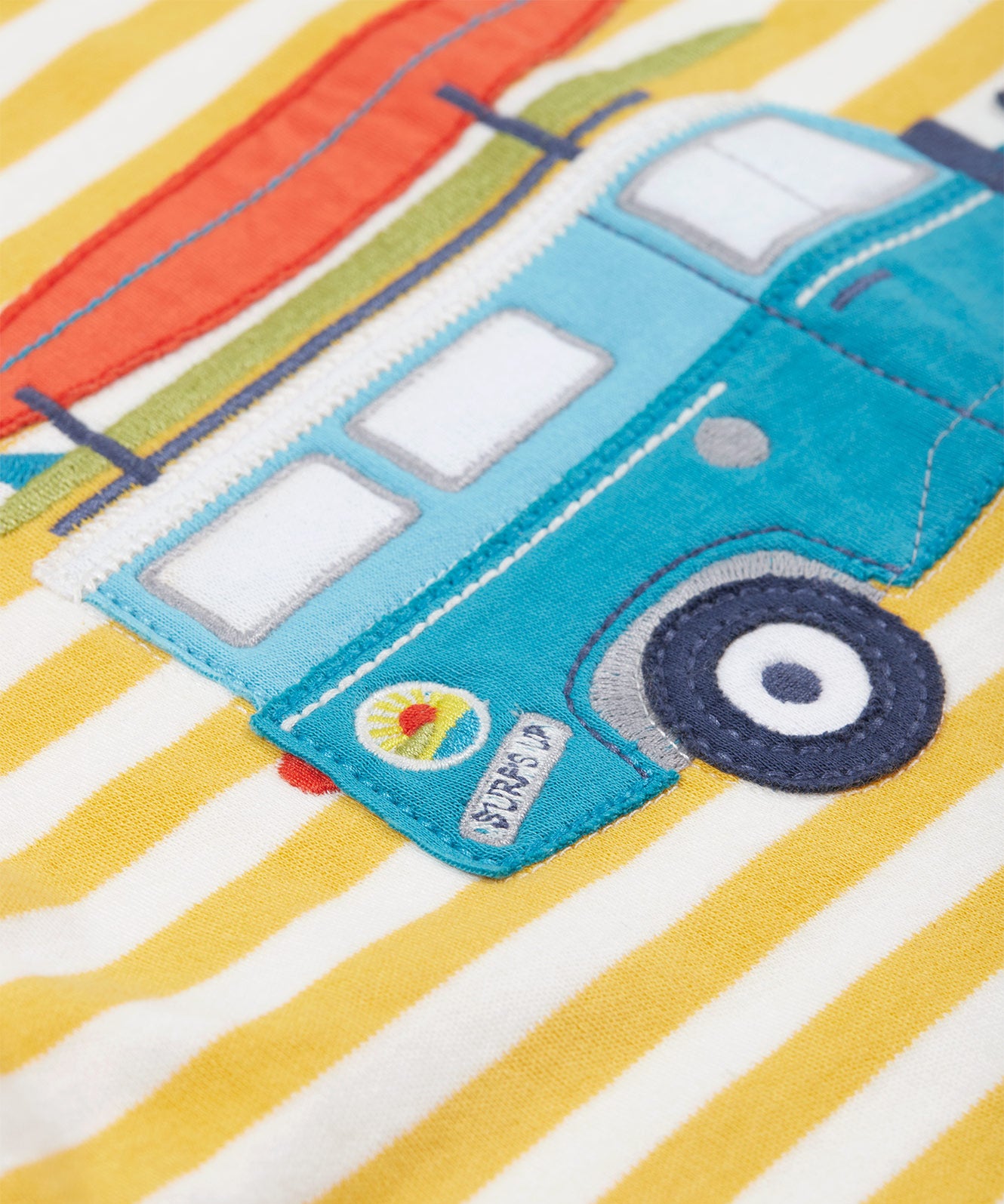 Close up of the Frugi blue camper van appliqué on the Frug kids tshirt. showing the decal detail on the campervan on yellow and white stripe organic cotton fabric