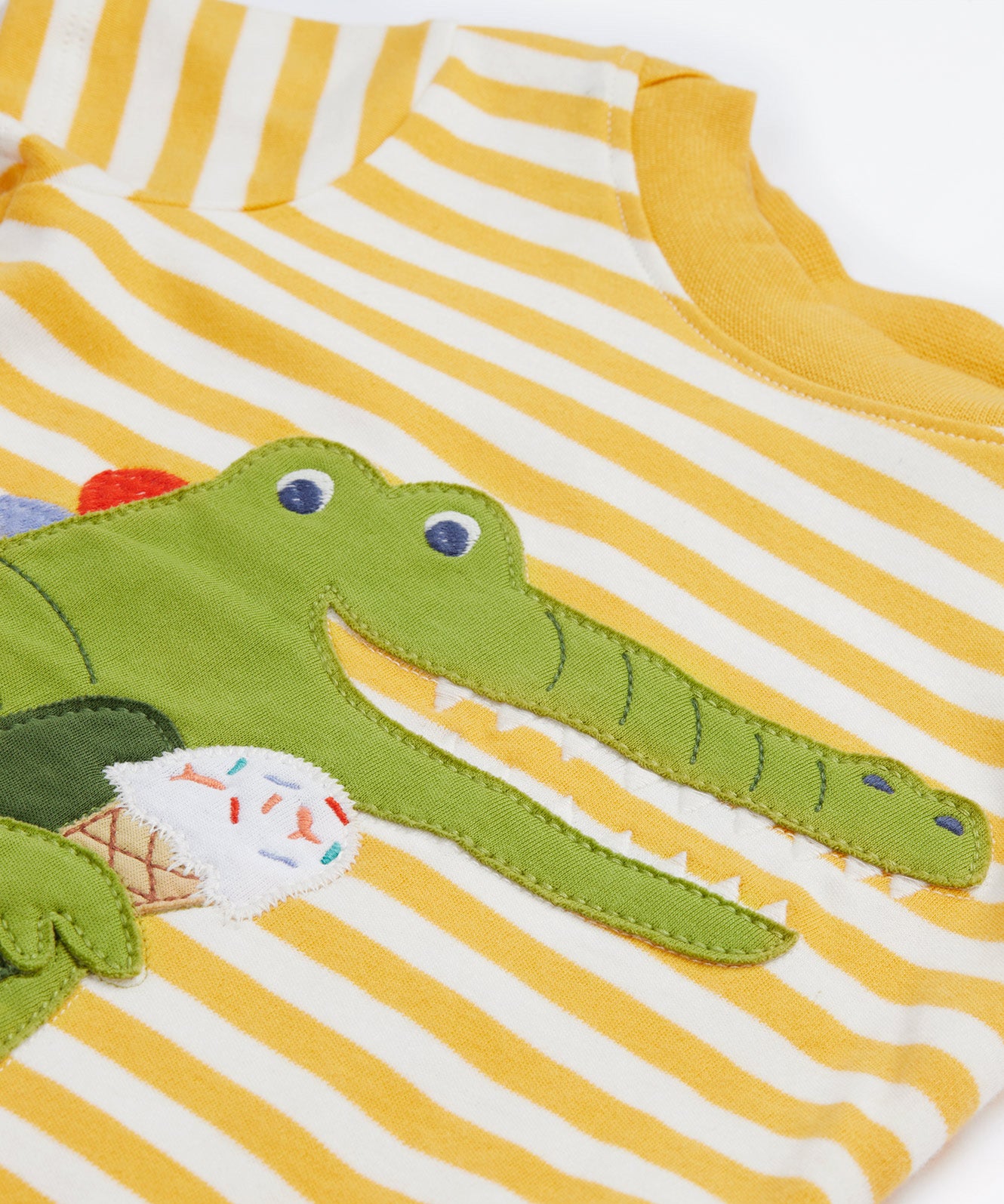 A close up of the Frugi kids wrap around crocodile outfit. Showing the front of the t shirt with crocodile eating a ice cream applique on yellow and white stripe organic cotton fabric