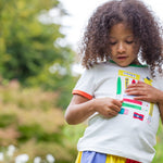 A child standing outside wearing the Frugi Xavier t-shirt. White with different coloured appliqués of art tools. The child is also wearing the Frugi rainbow jersey skirt.