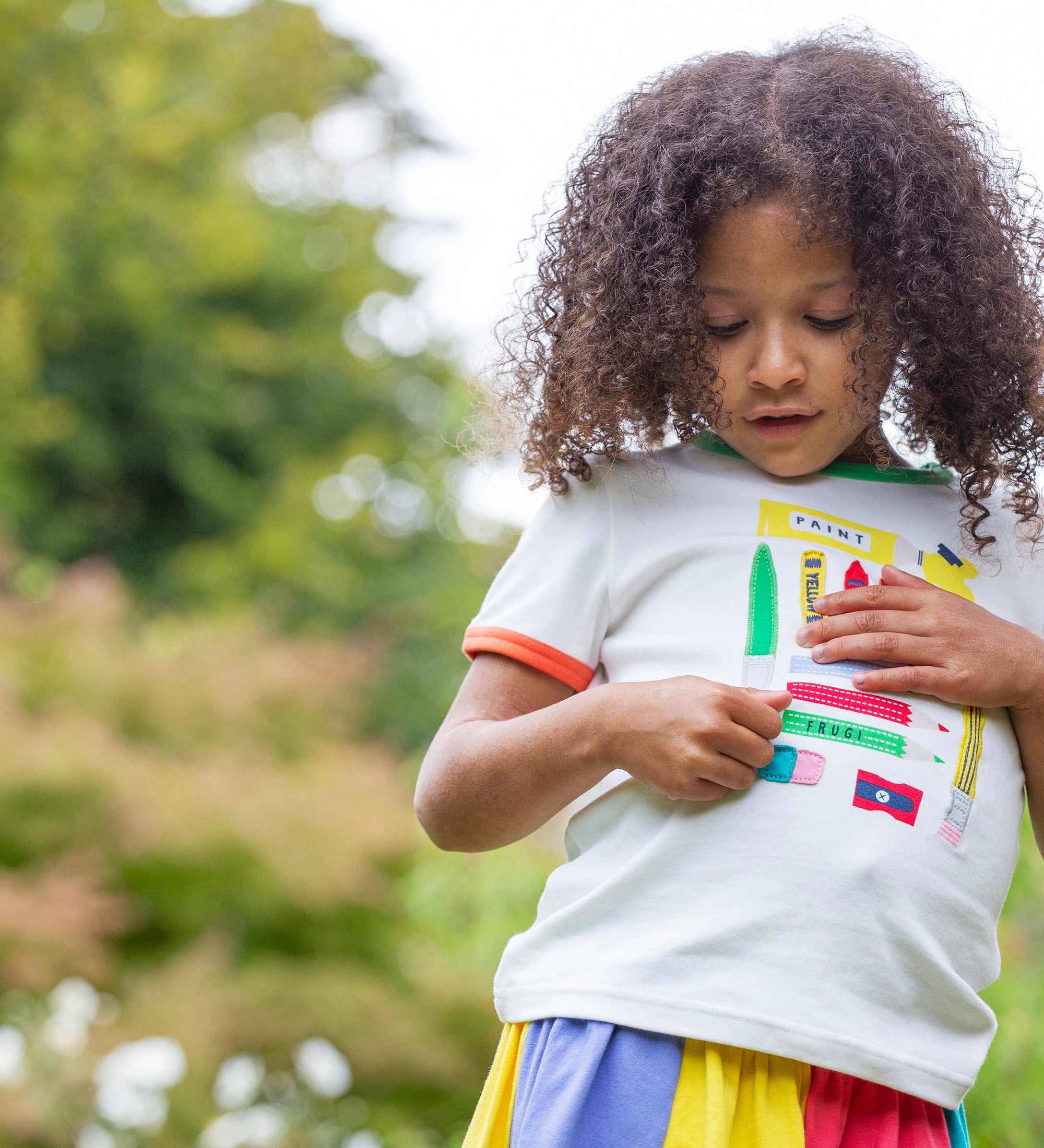 A child standing outside wearing the Frugi Xavier t-shirt. White with different coloured appliqués of art tools. The child is also wearing the Frugi rainbow jersey skirt.