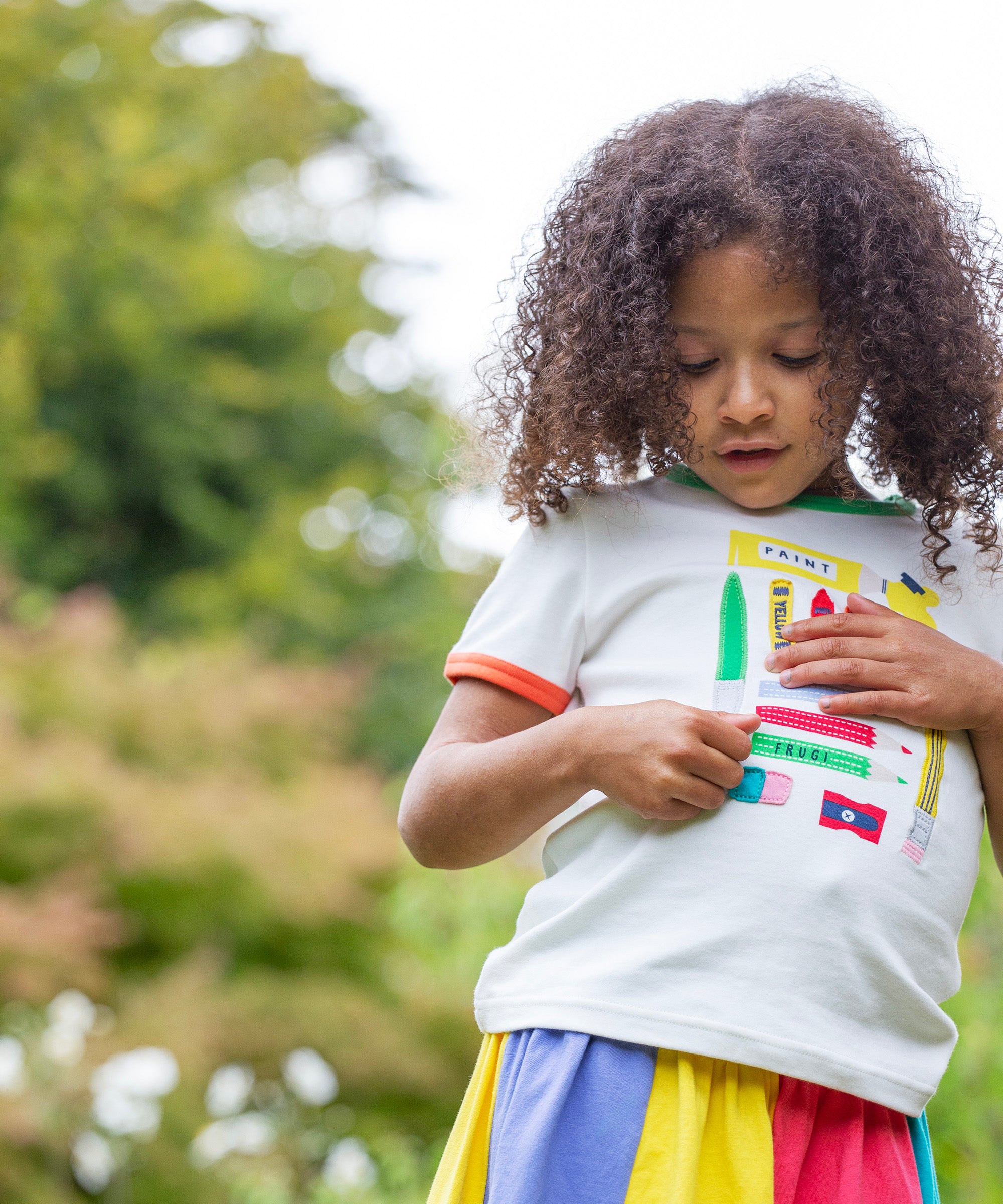 A child standing outside wearing the Frugi Xavier t-shirt. White with different coloured appliqués of art tools. The child is also wearing the Frugi rainbow jersey skirt.