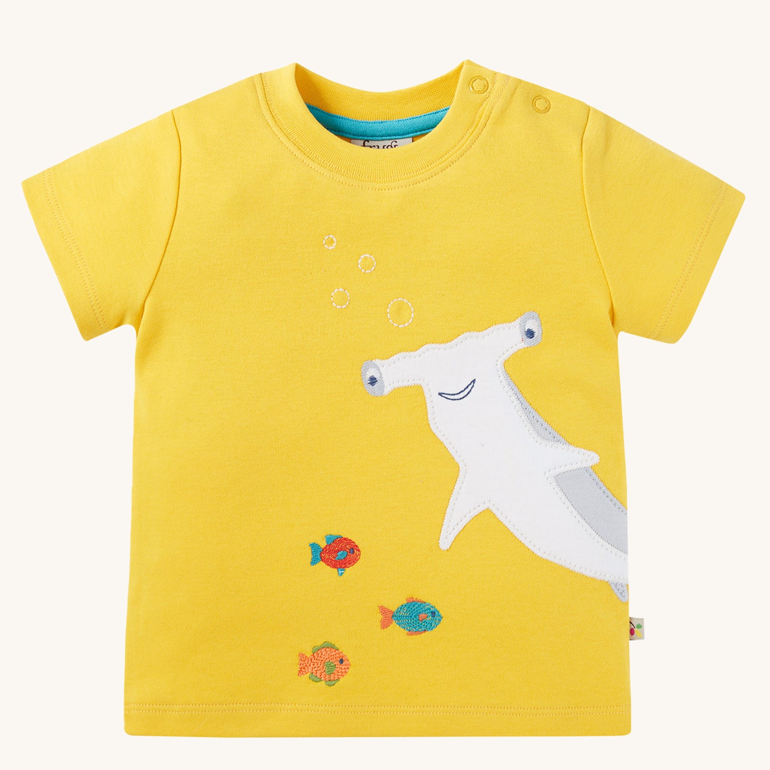 Frugi Children's Organic Cotton Jaime Applique T-Shirt - Shark. The front of the short-sleeved t-shirt showing shoulder pop fasteners, a fun Hammerhead Shark applique design and colourful fish on a sunny yellow fabric. T-shirt is on a cream background