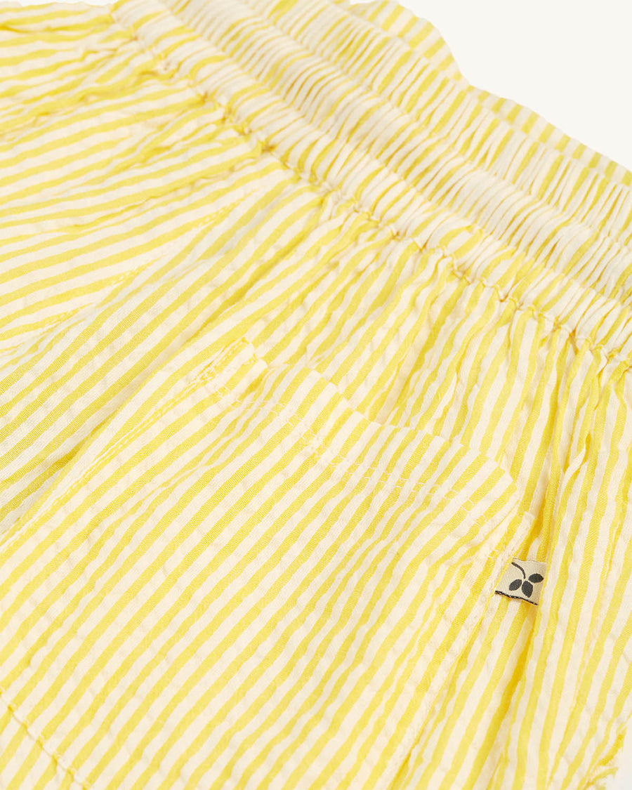 A child happily standing in a green house with their hands in the pockets of the Frugi Archie Seersucker Shorts - Dandelion. A bright yellow and white striped short with an elasticated waist