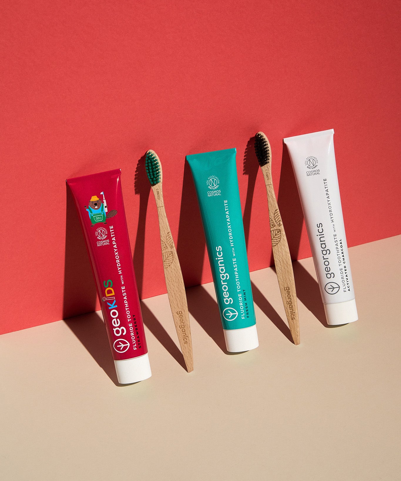 Georganics strawberry, activated charcoal and fresh mint toothpaste tubes standing next to two wooden Georganics toothbrushes on a neutral floor and red background