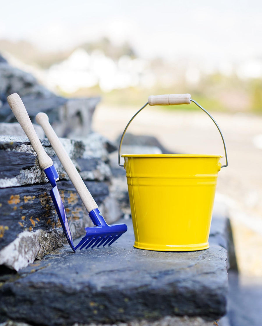 A yellow Glucksakfer bucket pictured outdoors on slate steps with a blue metal spade and rake next to them