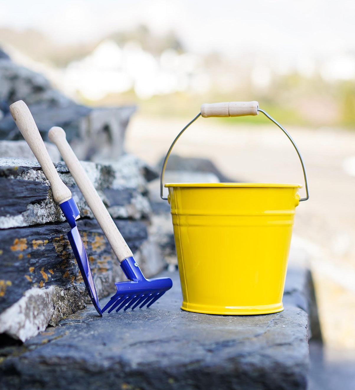 A yellow Glucksakfer bucket pictured outdoors on slate steps with a blue metal spade and rake next to them