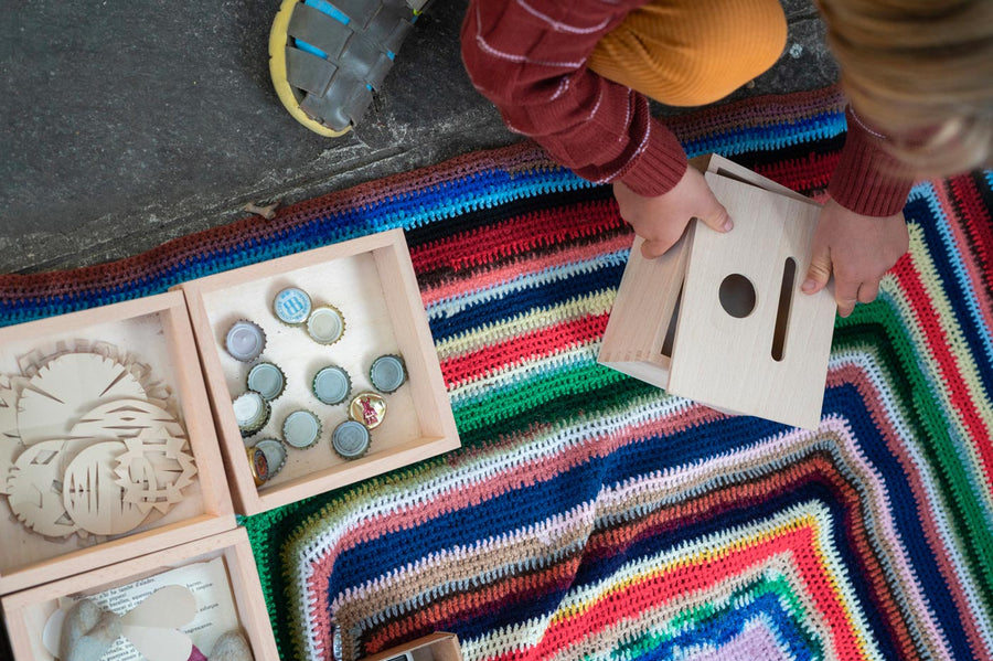 Close up of child crouched down next to some Grapat wooden boxes filled with bottle tops and paper shapes