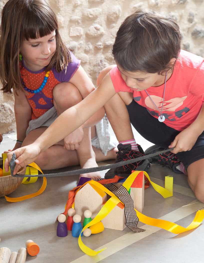 Grapat Six Wooden Houses Set in different simple geometric shapes in natural wood and colourful painted roofs. Two children playing with the houses in a village scene with colourful peg people and draping ribbons over the houses. Stone wall background.