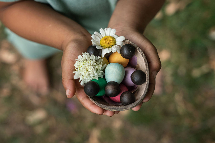 Close up of hands holding the Grapat dark baby nin peg doll toys in a wooden bowl with a white flower