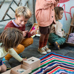 Children crouched down on a rainbow striped blanket around the Grapat plastic-free wooden permanence box