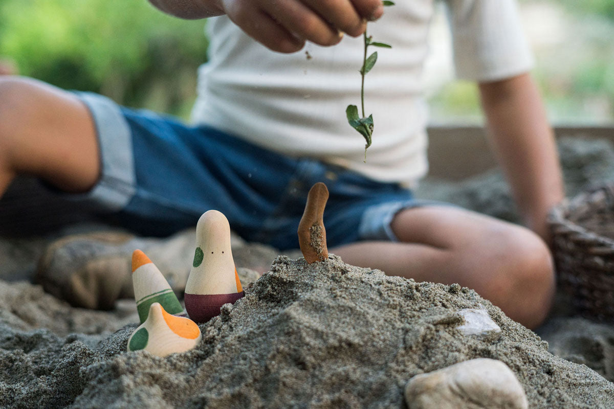 Close up of a hand holding a green plant stem above some sand with the Grapat wooden ooh lala figures sticking out