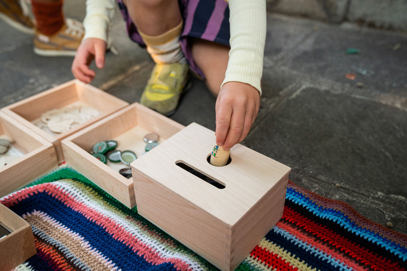 Close up of child's hand putting a Grapat nin inside the Grapat wooden permanence box on a striped blanket