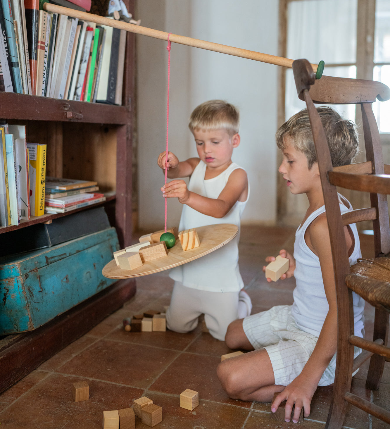 2 children sitting on a terracotta floor playing with the Grapat wooden pendulum set resting on a wooden chair and bookshelf with wooden blocks surrounding them and on the pendulum.