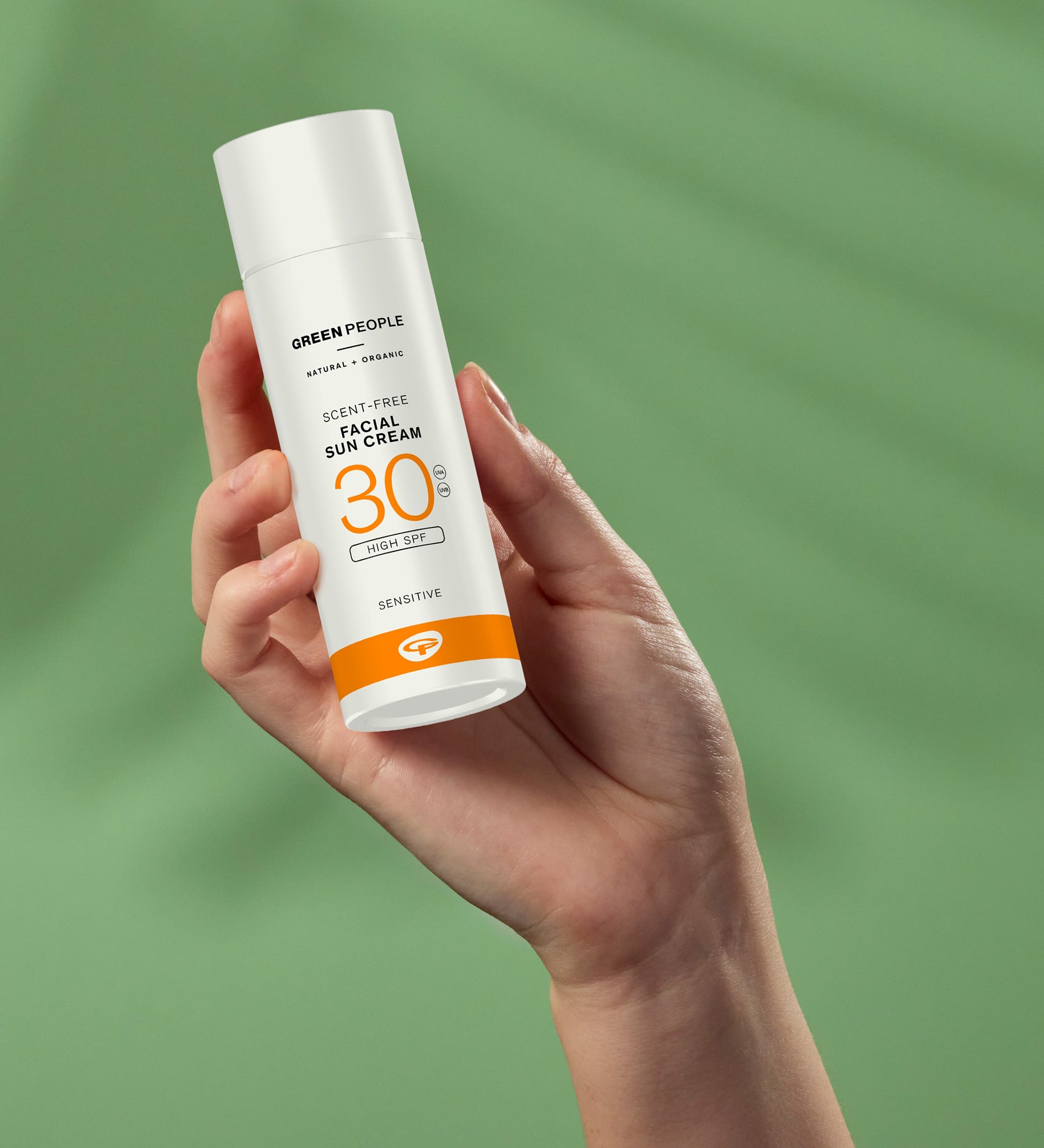 A hand holding up a tube Green People Facial Sun Cream SPF30 50ml against a green background. 