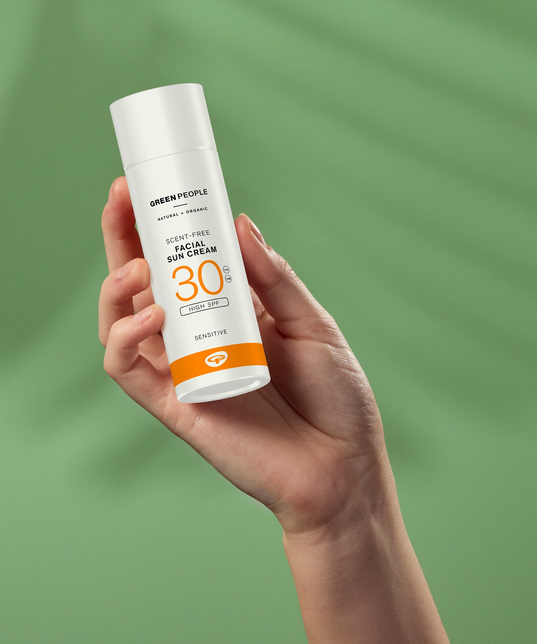 A hand holding up a tube Green People Facial Sun Cream SPF30 50ml against a green background. 