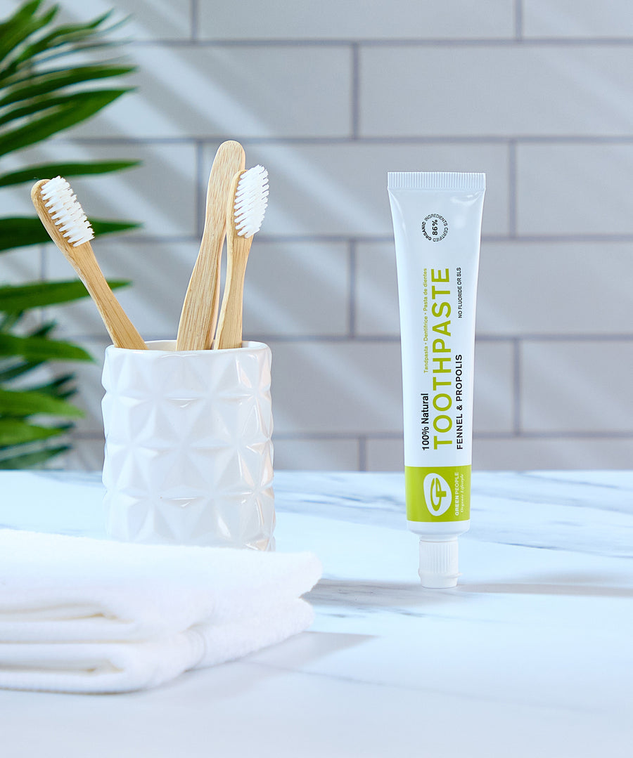 Green People Fennel & Propolis Natural Toothpaste on a marble surface in a bathroom next to a ceramic pot with bamboo toothbrushes.