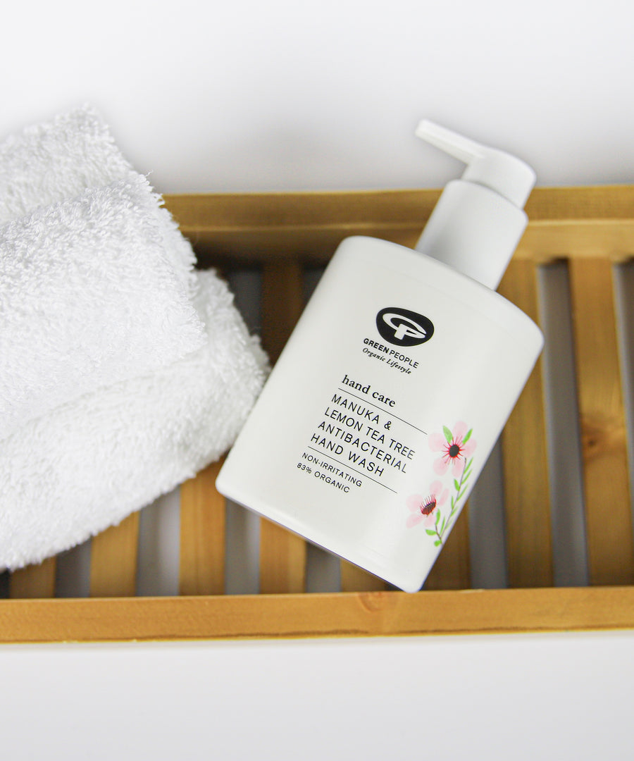 A bottle of Green People Manuka & Lemon Tea Tree Hand Wash placed on a bamboo bath shelf next to a fluffy white towel. 