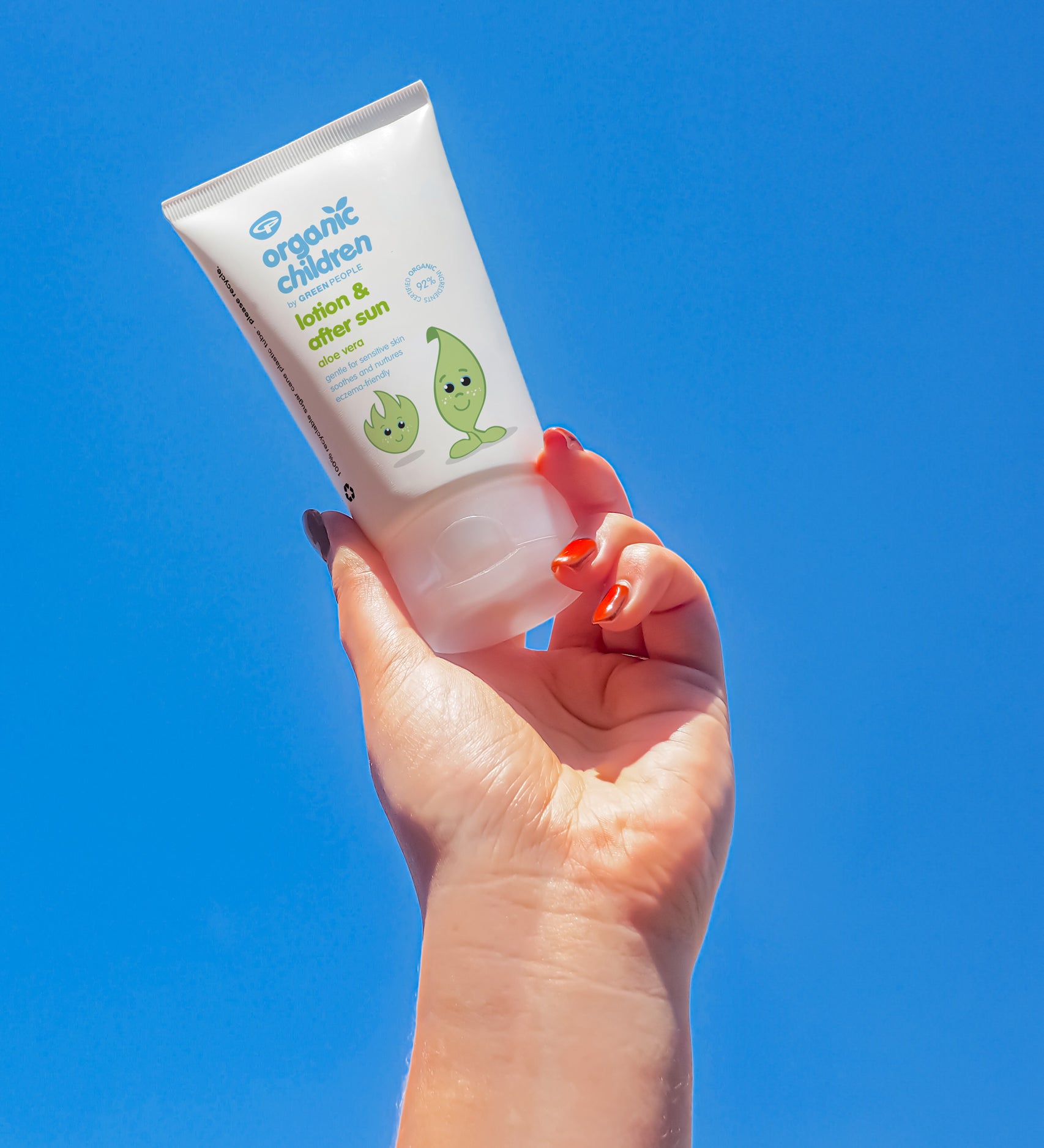 An adult's hand holding up a tube of Green People Children's Aloe Vera Aftersun Lotion against a clear blue sky. 