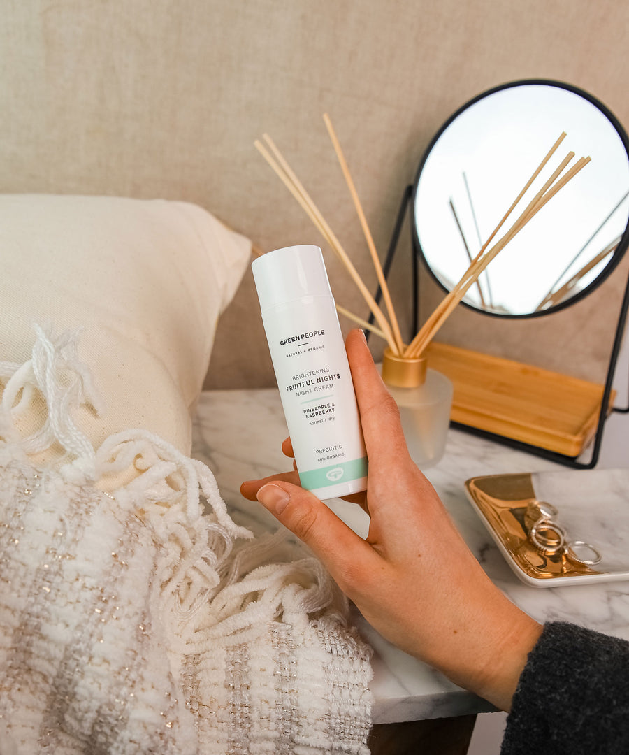 A person holding a bottle of Green People Fruitful Nights Night Cream in their hand. The cream is being held above a dressing table. 