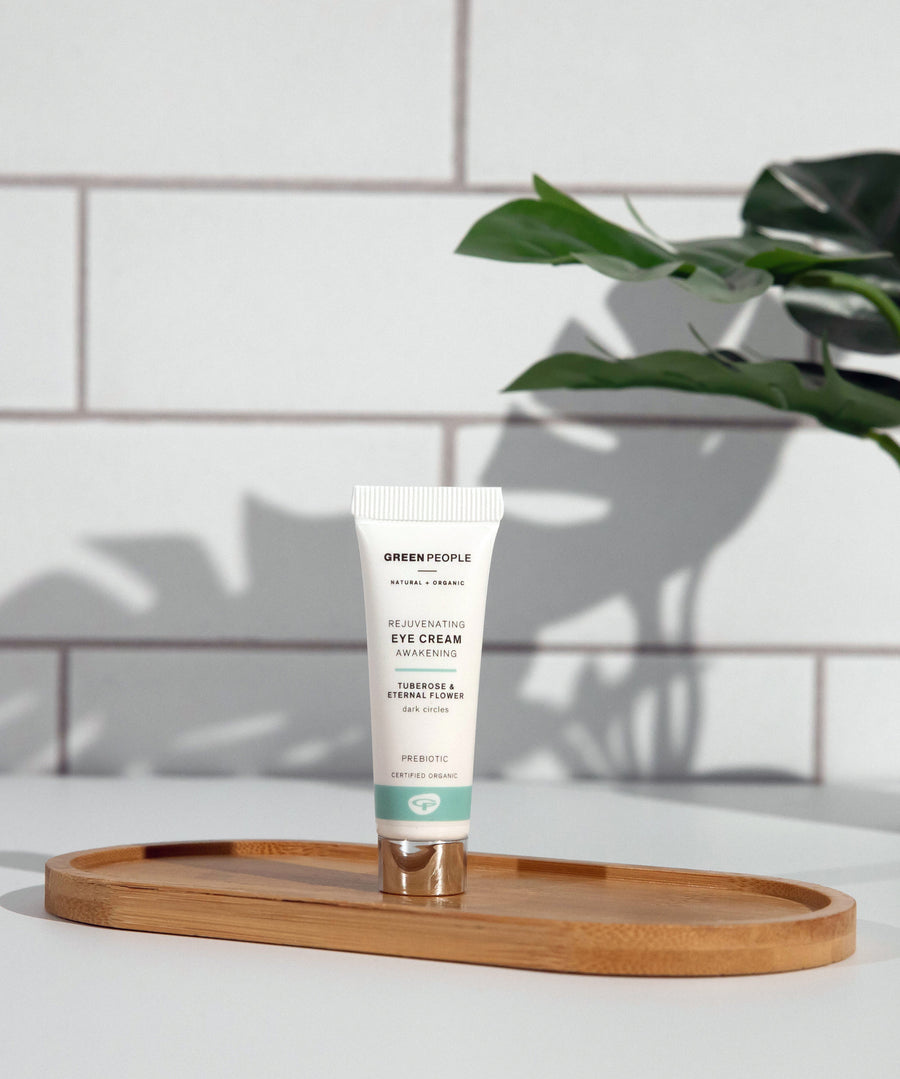 A tube of Green People Rejuvenating Eye Cream placed on an oval shaped bamboo tray in a bathroom. 