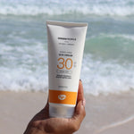 A tube of Green People Sun Cream SPF30 being held up in a person's hand. The sea can be seen in the background. 