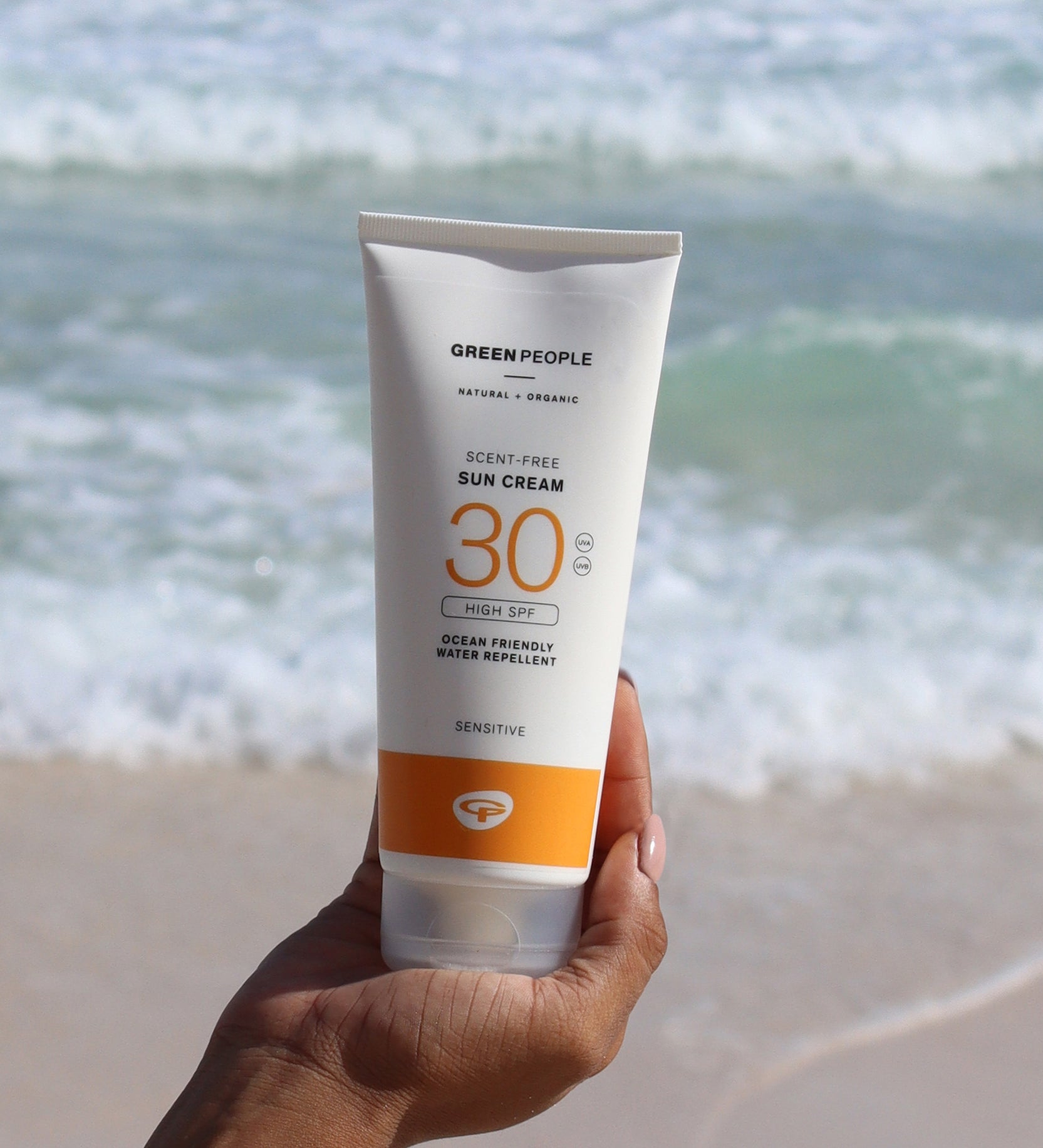 A tube of Green People Sun Cream SPF30 being held up in a person's hand. The sea can be seen in the background. 