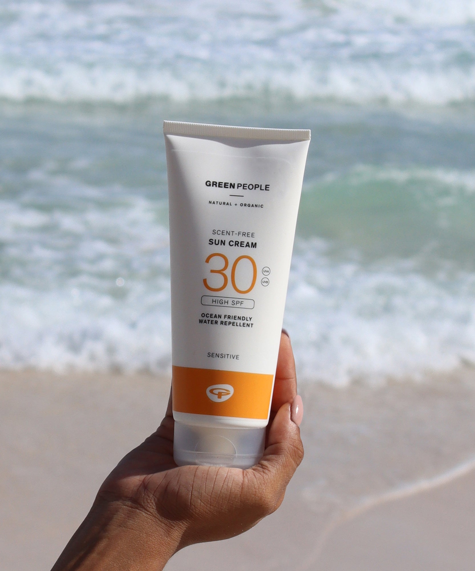 A tube of Green People Sun Cream SPF30 being held up in a person's hand. The sea can be seen in the background. 