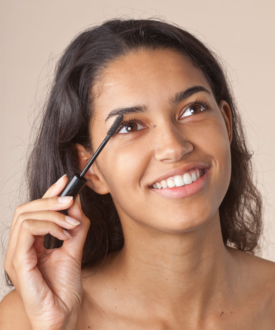 A person with dark hair applying the Green People Volumising Black Mascara to their eyelashes. The person is looking up and smiling. 