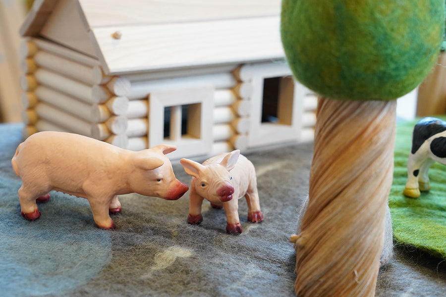 Close up of the green rubber toys pig and piglet figures on some grey felt in front of a small wooden hut toy