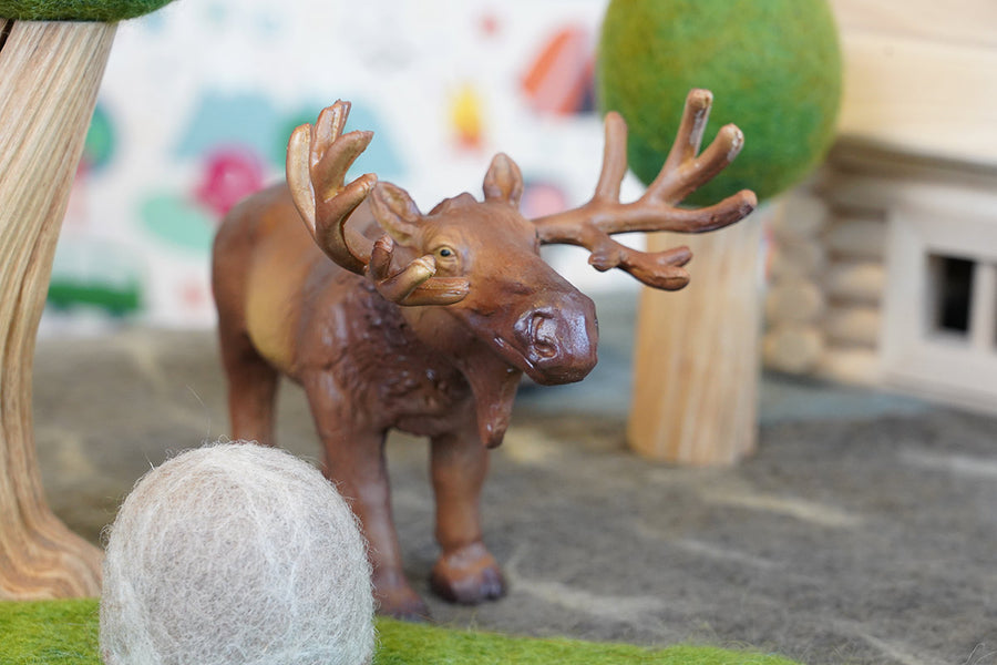 Close up of a green rubber toys sustainable moose figure on some grey felt in front of a papoose wooden tree