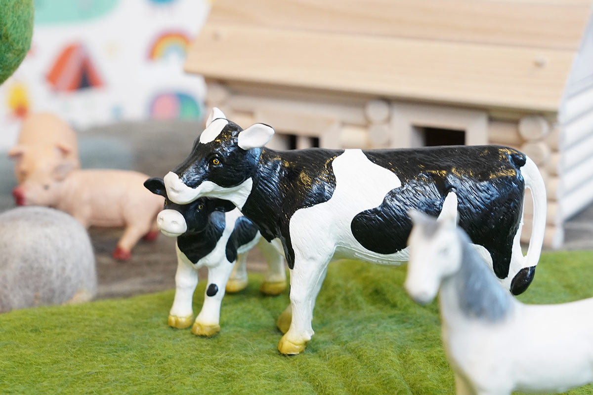 Close up of the green rubber toys cow figures stood on some green felt in front of a wooden house toy