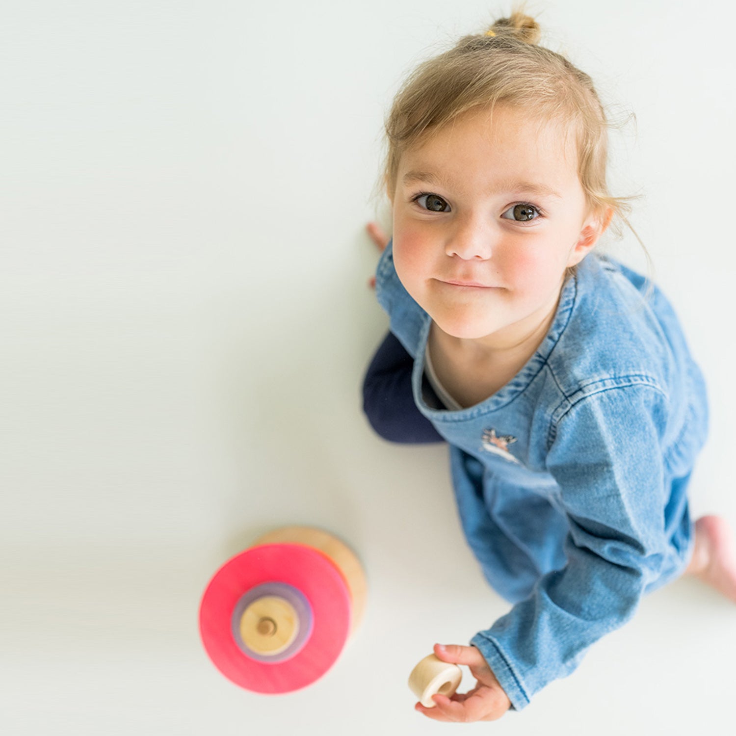 Grimm's Small Conical Stacking Tower - Neon Pink - On a plain blue background. A child looks up towards the camera whilst playing with the toy.