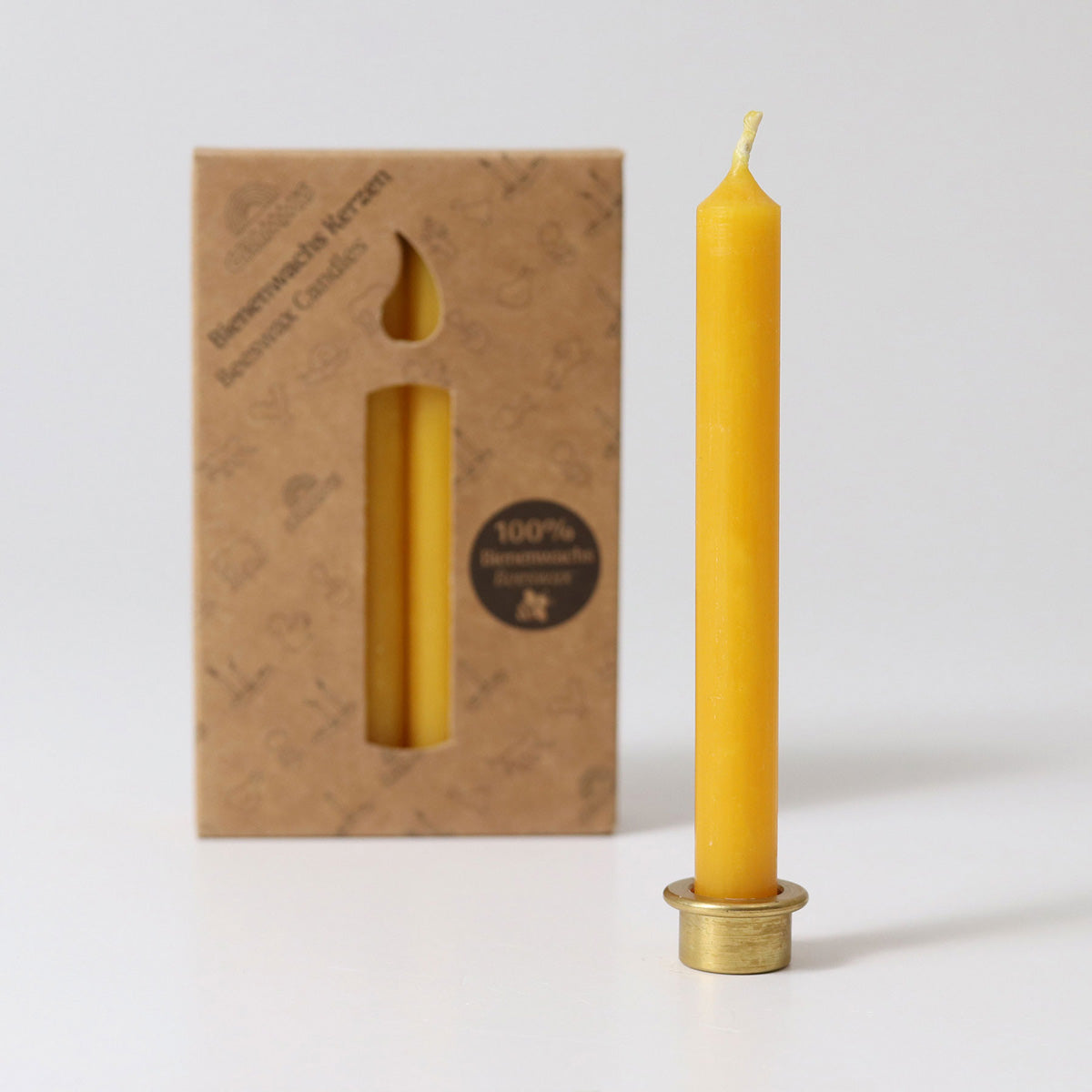 A pack of 12 Grimm's amber 100% beeswax candles in a cardboard box with one candles at the front in a brass holder. White background.