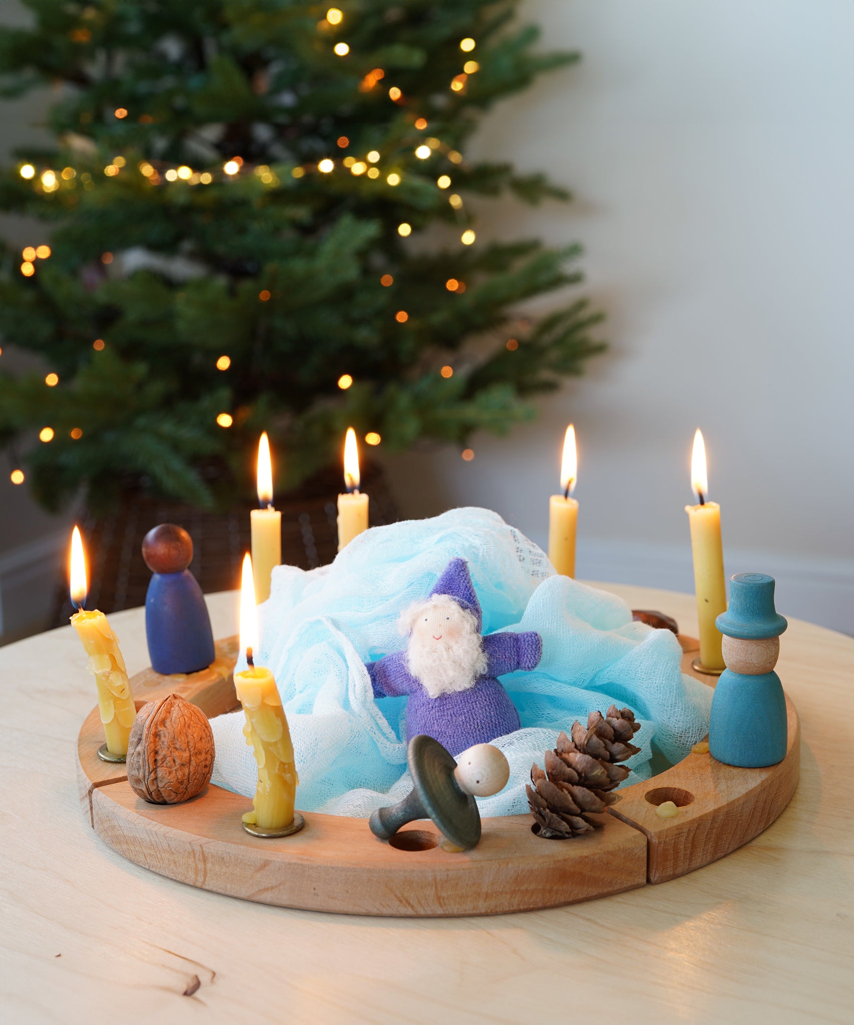 A Grimm's 16-Hole Natural Celebration Ring decorated in a Winter theme with candles, Nins, Magos, Grimm's dwarfs, pine cones. A Christmas tree with twinkling fairy lights can be seen in the background. 