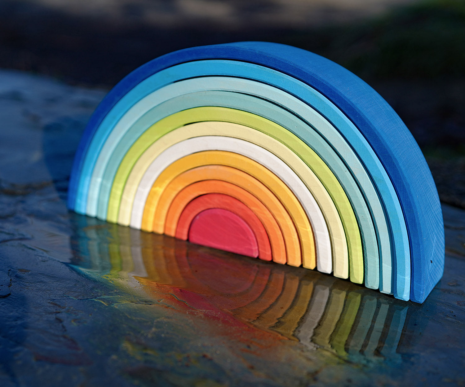 The Grimm's Gwawr Sunrise 12 Piece Rainbow shown on a wet slate surface outdoors. The reflection of the rainbow can be seen in the water,.