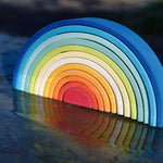 The Grimm's Gwawr Sunrise 12 Piece Rainbow shown on a wet slate surface outdoors. The reflection of the rainbow can be seen in the water,.