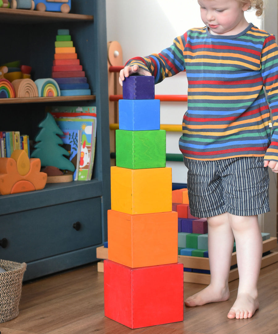 A child playing with the Grimm's Large Coloured Boxes Set. The boxes are all stacked up on top of each other, the child is placing the smallest block on the top