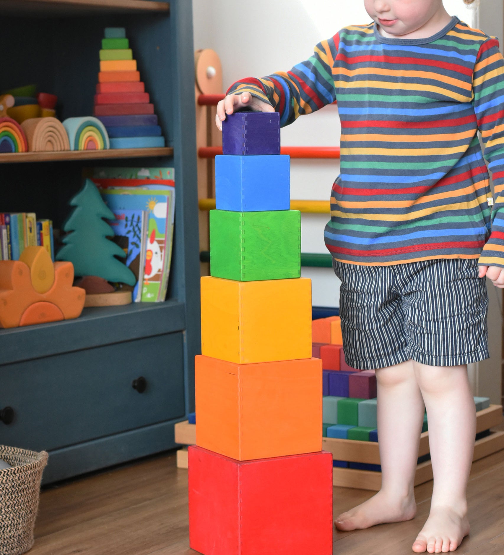 A child playing with the Grimm's Large Coloured Boxes Set. The boxes are all stacked up on top of each other, the child is placing the smallest block on the top