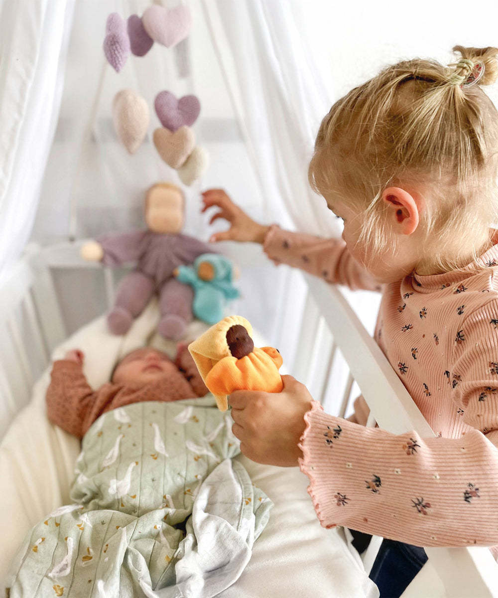 An older child is looking over the side of a white cot with a baby inside. The older child is holding the Grimm's Lavender Doll Sun in their hand. The image also shows other Grimm's Doll at the head of the cot with a pink and purple fabric heart mobile above the cot