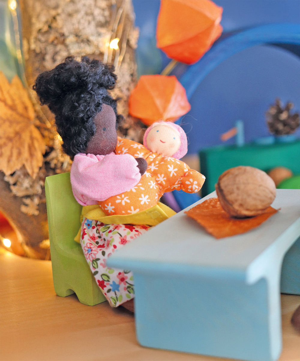 Grimm's Mrs Ebony is holding Baby Mia, whilst sat on a green wooden chair. The background has an autumnal theme with brown and orange colours, leaves and pinecones