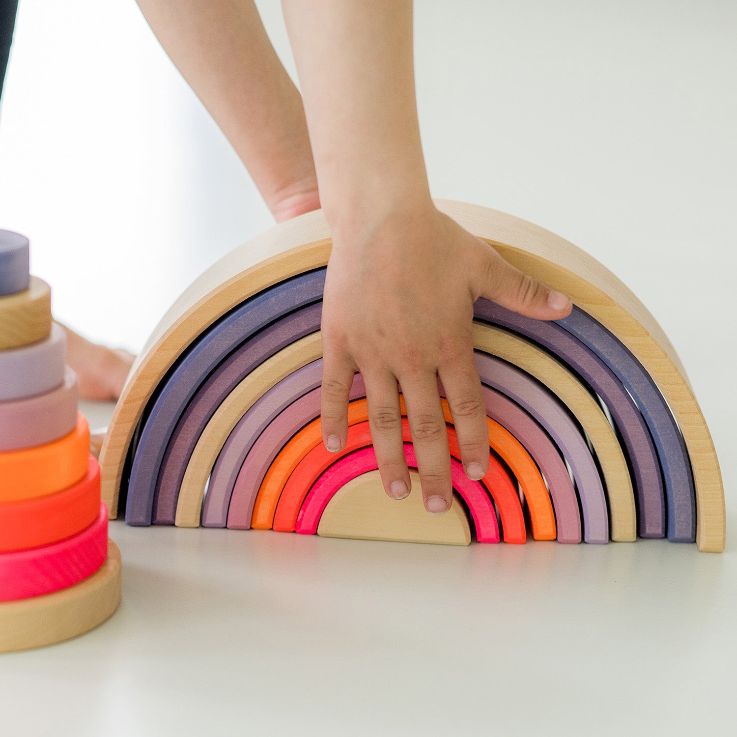 Grimm's 10-Piece Rainbow - Neon Pink. A child is leaning over the toy about to pick it up.