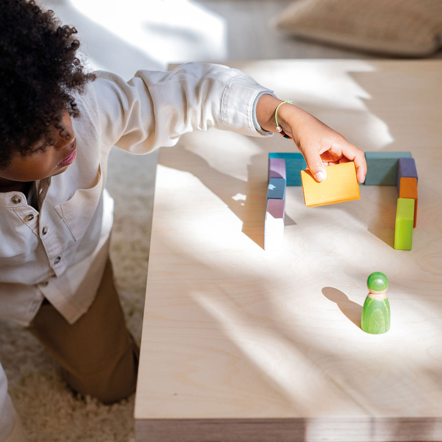 A child happily playing with the Grimms Wooden Pastel Duo Blocks. The child is in the process of building with a Grimms friend watching on