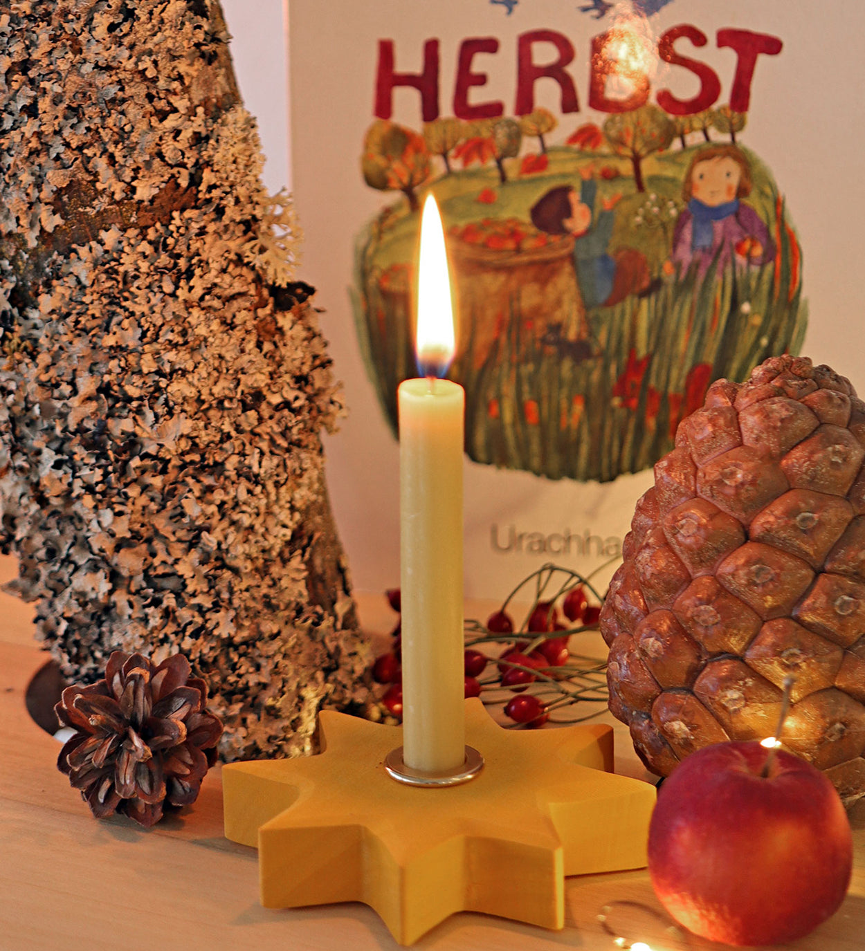 Grimm's Yellow Lifelight Star with a lit candle set up in a little seasonal display featuring a small apple and pine cones. 